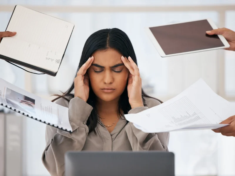 A woman at a laptop holding her temples as people present notebooks and paperwork.